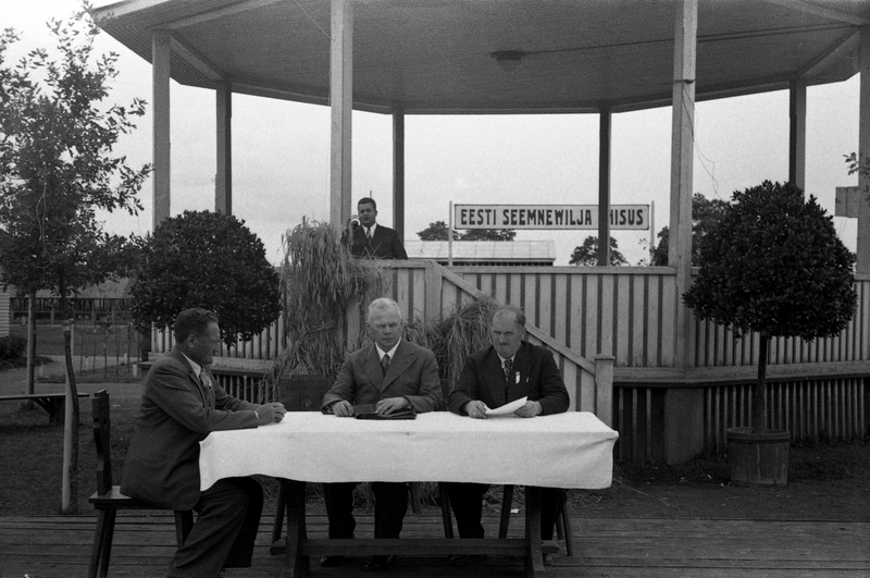 Kolm meest paviljoni ees laua taga istumas ja mees paviljonis kõnelemas