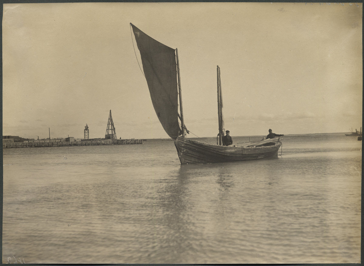 Vene-Balti laevatehas. Esiplaanil meri, millel kahemastiline purjepaat, kus kaks meest ja lauakoorem. Tagaplaanil  kraanad, ellingud ja tehasehooned