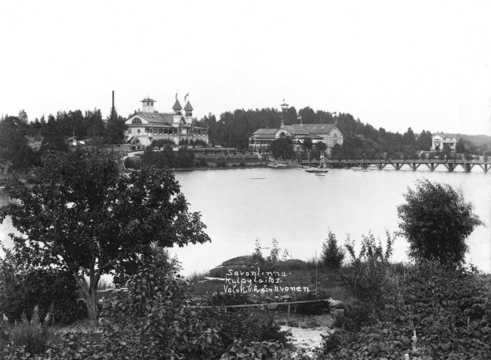 Kylpylaitos, Kasino juhlasaleineen ja Suruton-huvila Haapasalmen vastarannalta otetussa kuvassa. Taustalla Kylpylaitoksen näkötorni.	Baths, Casino and Villa Suruton in Savonlinna