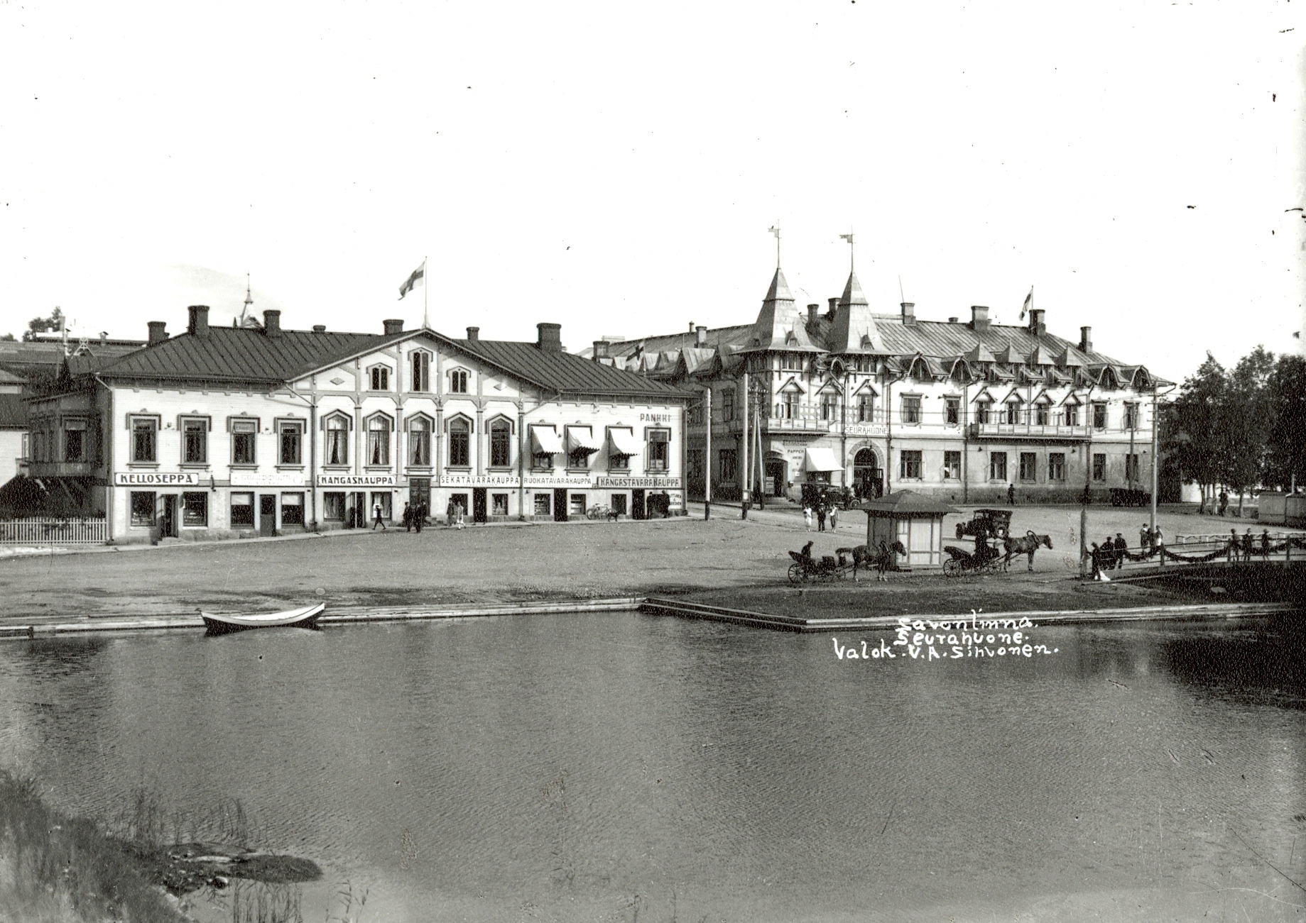 Miljonatalon rannasta yli Haapasalmen itään. Sikion talossa Suomen lippu, "Kelloseppä", "Kukka- ja siemenkauppa", "Kangaskauppa", "Sekatavarakauppa", "Ruokatavarakauppa", "Kangastavarakauppa" (Huttunen & Hirvonen), "Pankki". Seurahuone liputettu Suomen lipuin. kesä 1923. View east from Miljoonatalo