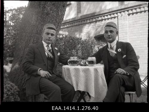 Plastic negative from two seated men. Two men sitting under a large tree behind a small table in cliffs and colored tapes. The right man has a cigarette between the buttons, the left tape has an unreadable text. The envelope is the same text as the previous one.