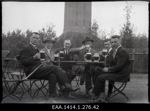 Plastic negative from the company in the beer table. Six men sit in the garden cafe and drink beer. Two men have summer cables in their heads. The rear appears a monument or an unknown building. The envelope is the same text as the previous one.
