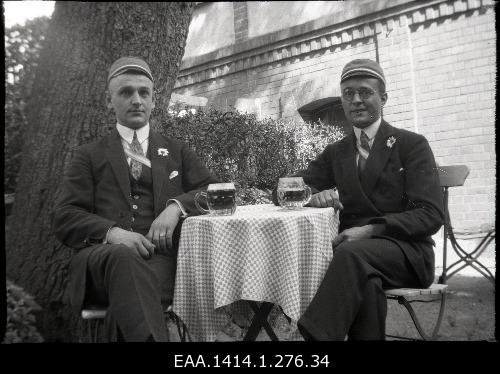 Plastic negative from two men. Two men in suits, and men with clothes and tapes sitting under large tree chairs. There are two round breweries on the table with a small square linen.The same text on the whole is the same as the previous one.