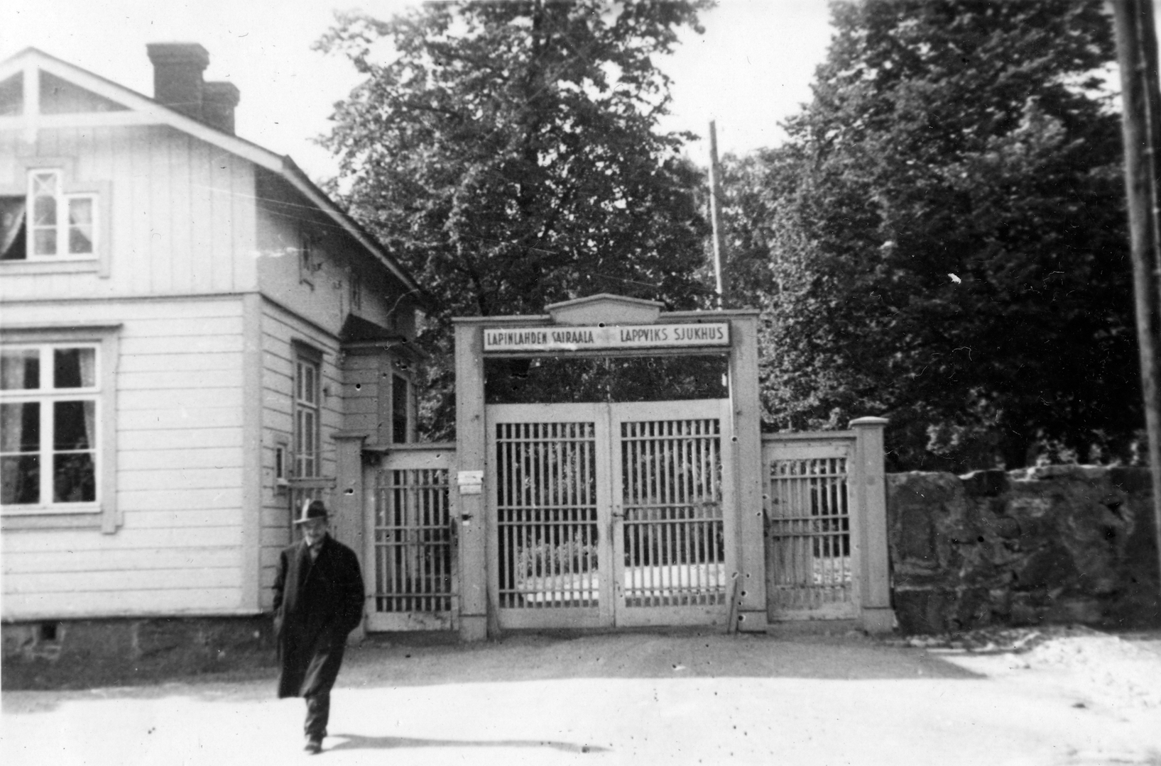 Lapinlahden sairaala-alueelle johtava portti ja portinvartijan tupa, joka on puinen asuinrakennus. Portin päällä on teksti "LAPINLAHDEN SAIRAALA LAPPVIKS SJUKHUS". Portin oikealla puolella on kivimuuri. Portin suunnasta kävelee hattupäinen mies. 1930-luku.