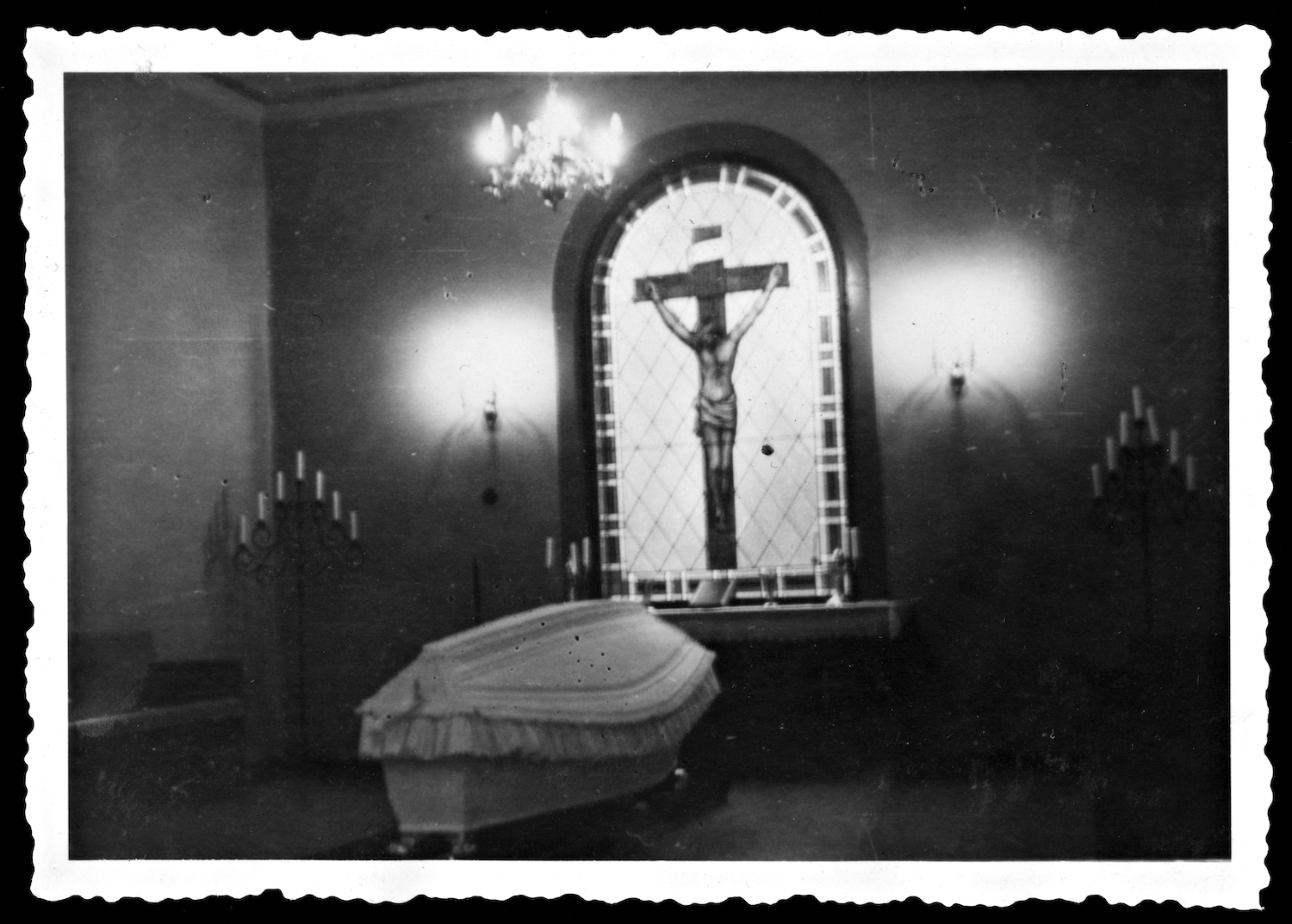 Lapinlahden sairaala. Valkoisella kangaspäällysteellä koristeltu ruumisarkku alttarin äärellä. Alttaritauluna on lasimaalaus ristille naulatusta Jeesuksesta. Alttarin molemmilla puolilla on lattialla seisovat seitsenhaaraiset kynttilänjalat. Kuvan alla on ruotsinkielinen teksti: kanske? 1930-luku.