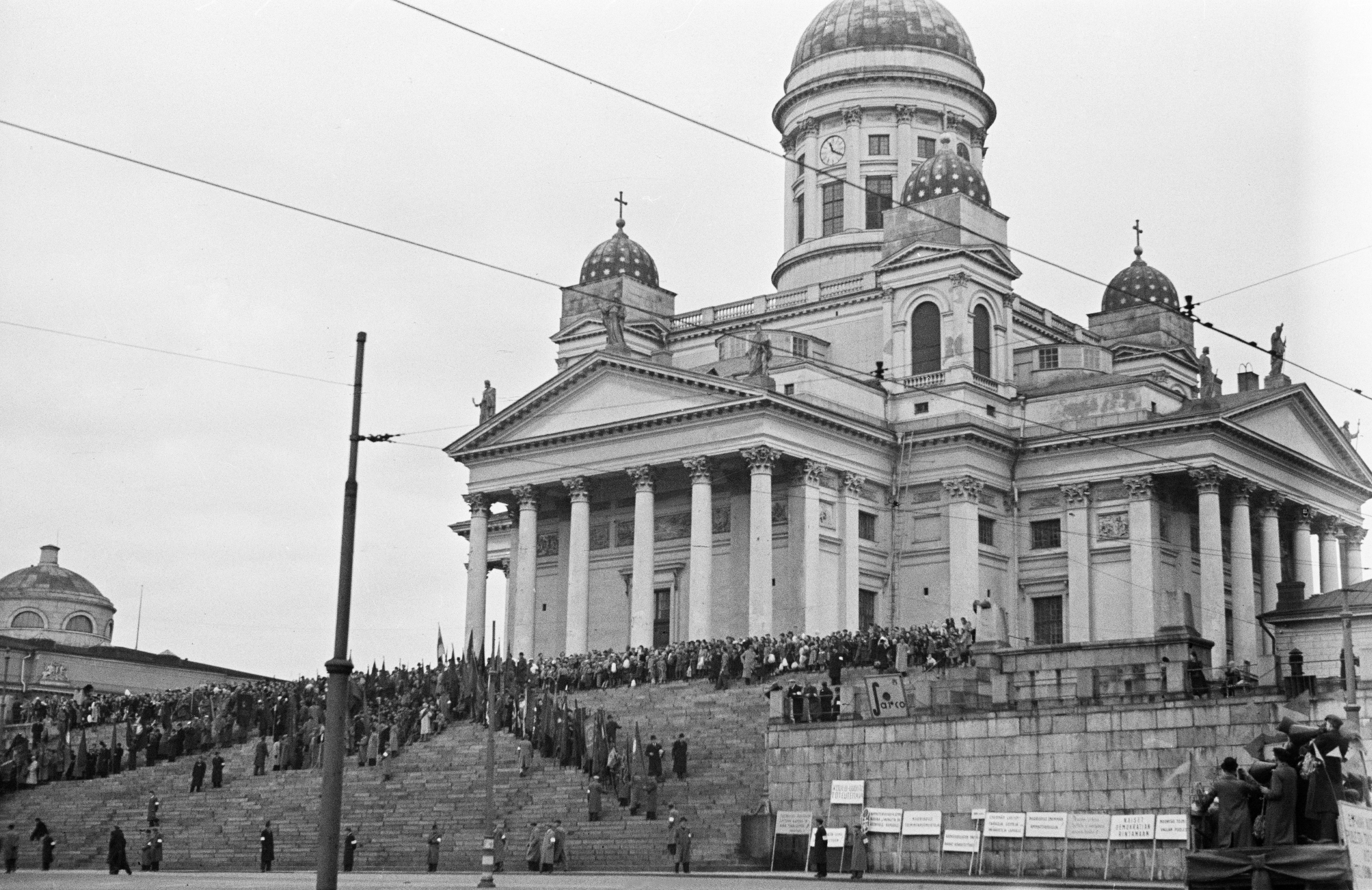 Vasemmiston yhteinen vappujuhla Senaatintorilla. Puhujakoroke ja lippurivistö Suurkirkon (=Helsingin tuomiokirkko) portailla.