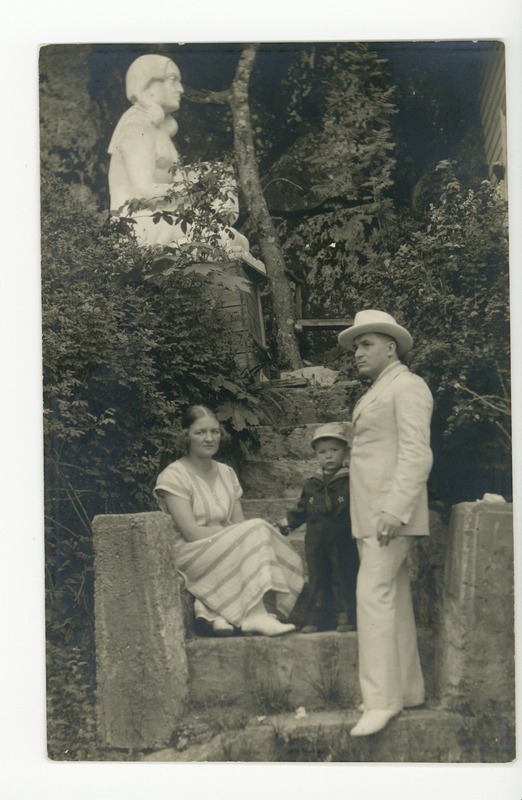 Kuju jalami astmel istuv vöödilises kleidis naine, seisavad väike madruseülikonnas poiss ja valges ülikonnas kaabus mees