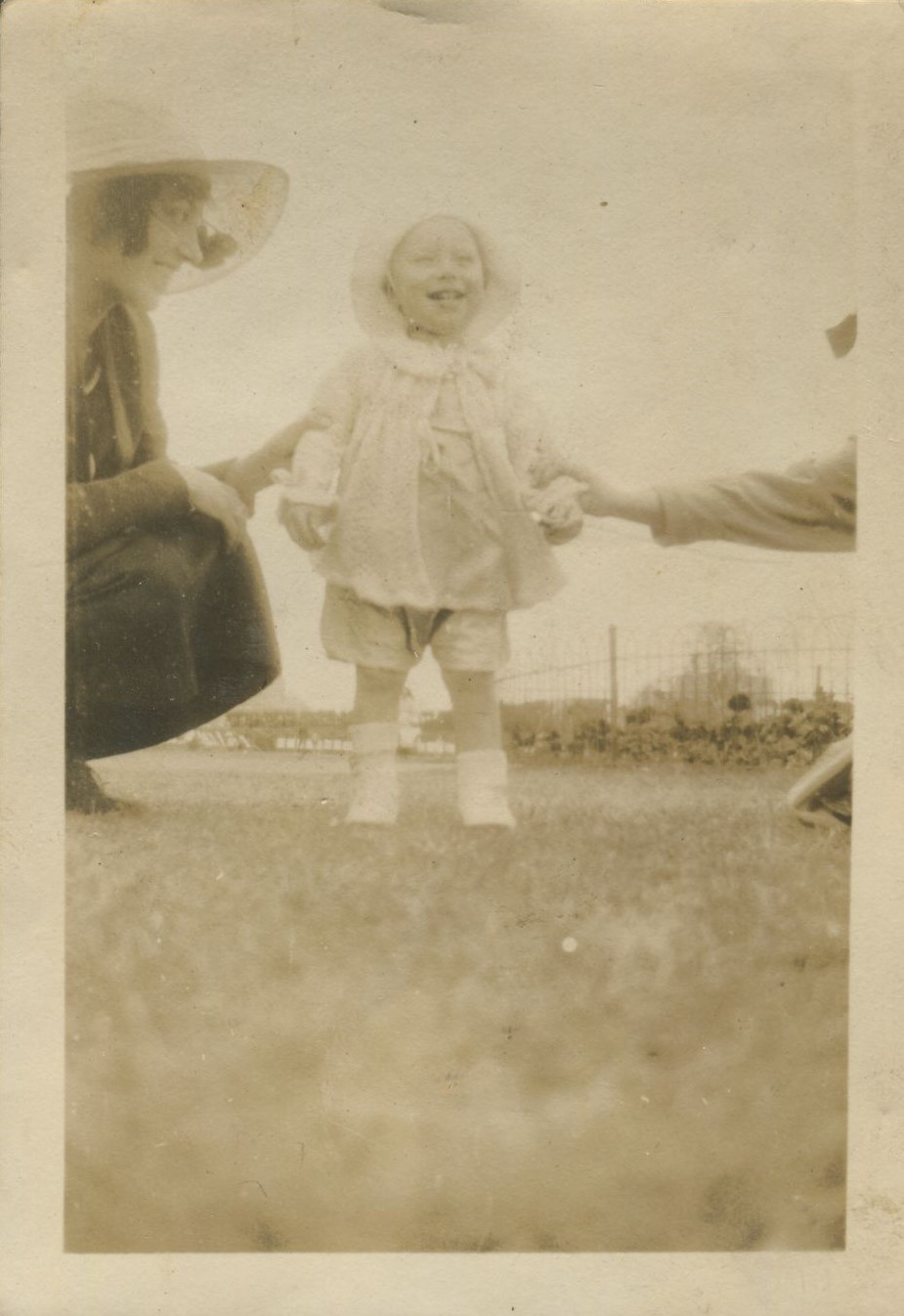 Toddler standing on the grass supported by two women