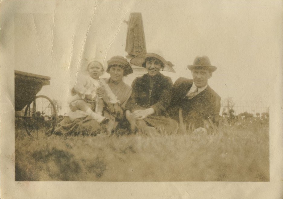 Family group seated on the ground, with a toddler and pram, in front of a statue