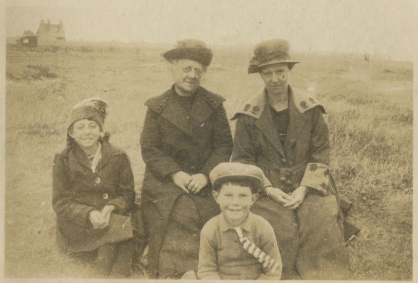 Two women and two children seated on the grass with house in distance