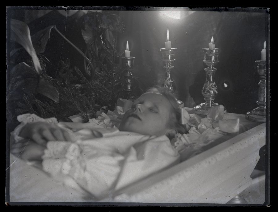 Matusefoto, laps avatud kirstus, kirstu ümbritsevad lilled ja küünlajalad.