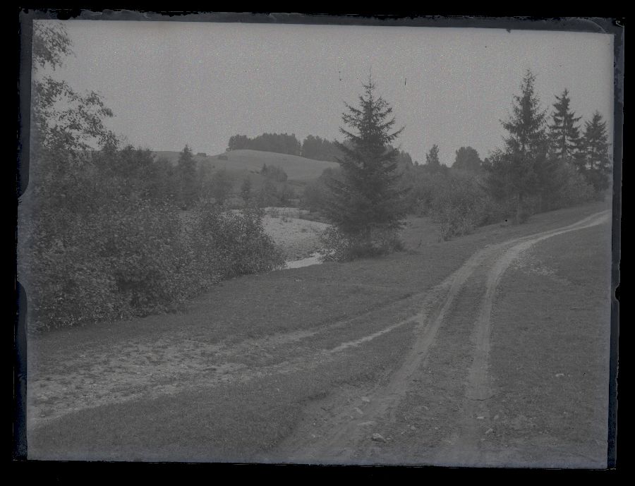 Maastikuvaade, paremat kätt kõrgel kaldapealsel mullatee, pildi keskel oja ja vasakul oja teine kallas küngastega.