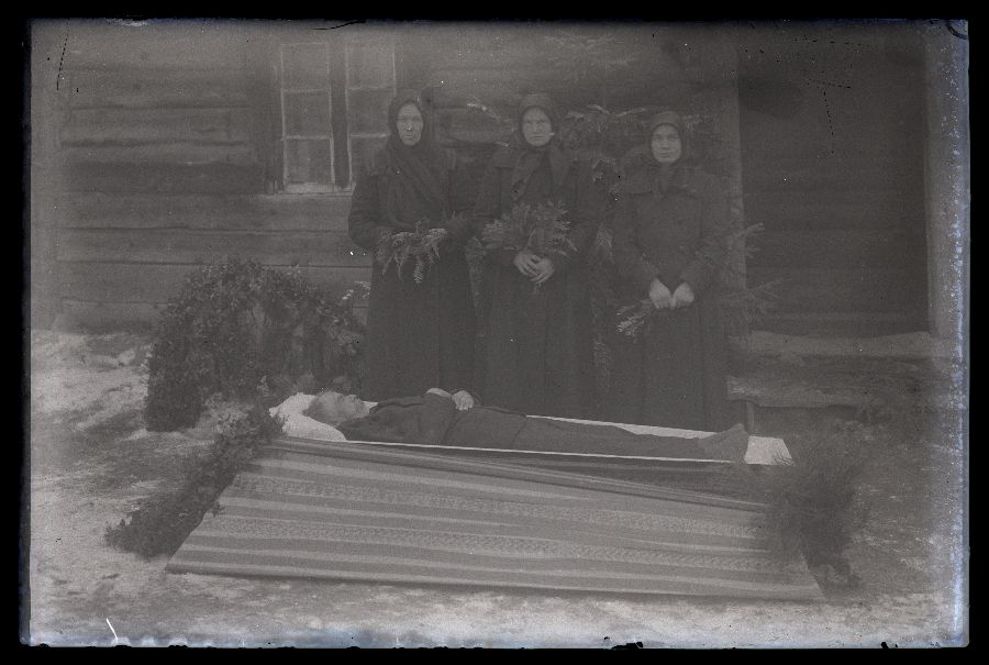 Matusefoto, vana mees avatud kirstus, kirstu taga seisavad kolm mustades riietes naist. Pildistatud taluhoone ees.