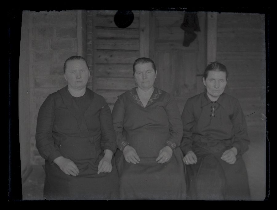 Grupifoto, kolm vana naist istuvad maja ees.