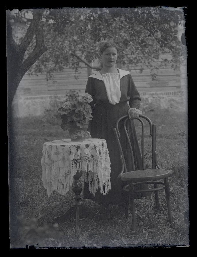 Naine aias, poseerib ümmarguse laua kõrval, millel potilill, taustal näha majasein.