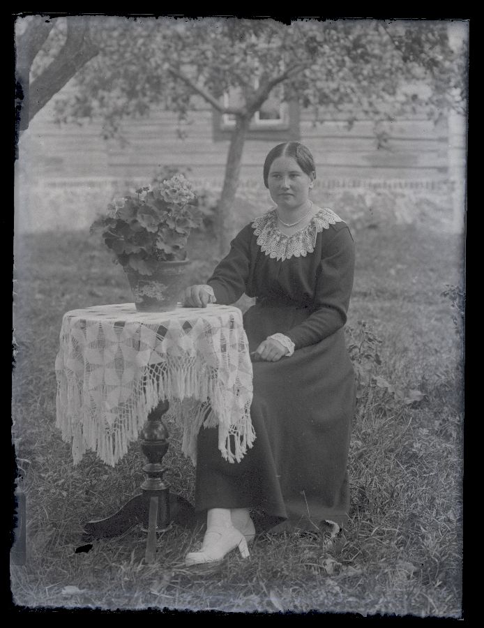 Naine aias, poseerib ümmarguse laua kõrval, millel potilill, taustal näha majasein.