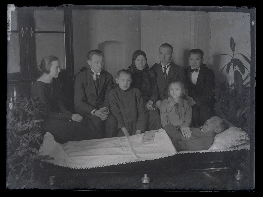 Matusefoto, vanem mees avatud kirstus, kirstu taga istuvad inimesed.