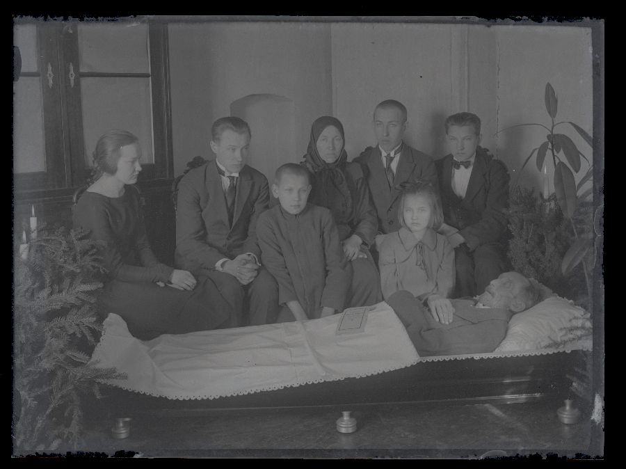 Matusefoto, vanem mees avatud kirstus, kirstu taga istuvad inimesed.