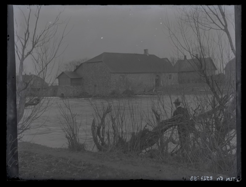Maastikuvaade vesiveskile kõrvalhoonetega, esiplaanil meesfiguur veekogu ääres, umbes 1910. aastast.