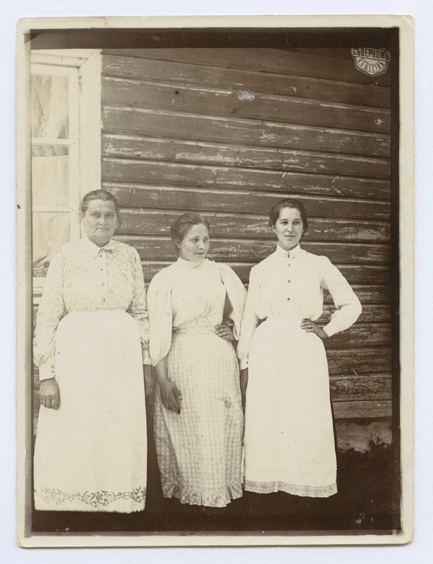 Kolm heledas riietuses teenija tüdrukut puumaja taustal.