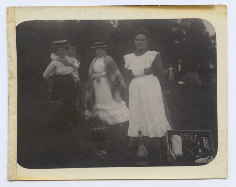 Väljasõit rohelisse. Esiplaanil valge põllega teenijatüdruk, tema ees priimus kastruliga, pudel ja kohver söögiriistadega.