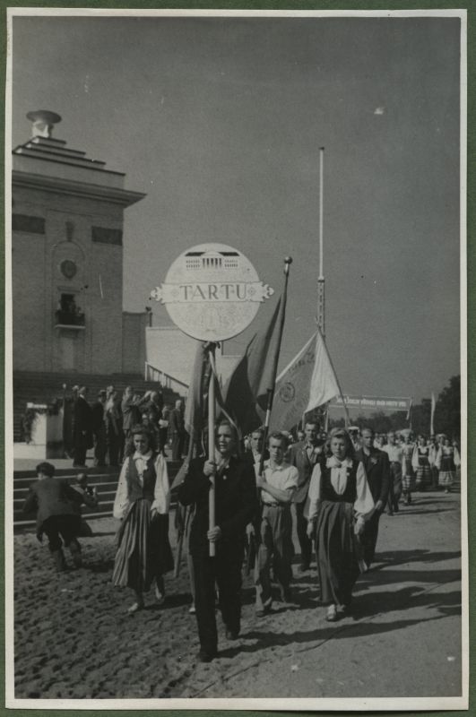 XII üldlaulupidu 28.-29. juunil 1947, Tallinnas.
12. üldlaulupeo osalejad rongkäigul jõudmas Tallinna lauluväljakule. Rahvariietes Tartu koorid jõudmas lauluväljakule. Ees kõnnib meesterahvas, kes kannab silti "Tartu", millel on näha Tartu Ülikooli peahoone kujutist. Tagapool järgnevad teisedki lipud ja kaugemal on näha loosungit. Paistab Tallinna vana laululava ja mõned pealtvaatajad.