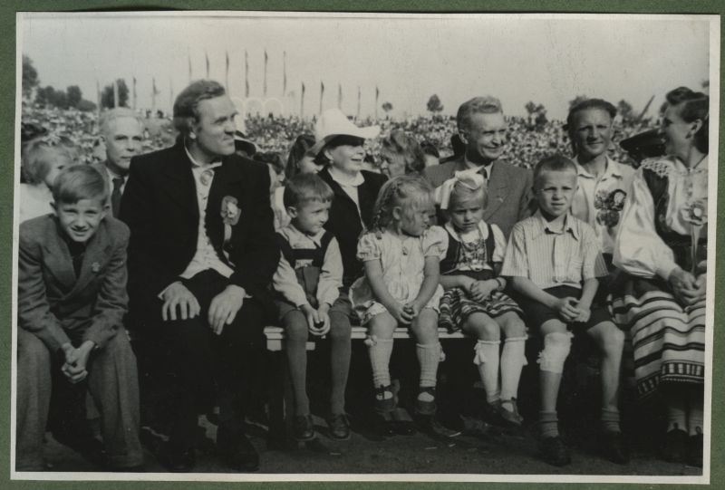 XII üldlaulupidu 28.-29. juunil 1947, Tallinnas.
Pealtvaatajad 12. üldlaulupeol Tallinna lauluväljakul. Pingil istuvad viist last ja rahvariietes paar. Taustal paistavad teisedki pealtvaatajad.