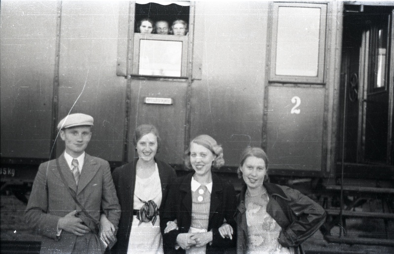 Kolm naist ja mees rongi ees seismas, tagaplaanil kolm inimest vaguniaknast välja vaatamas