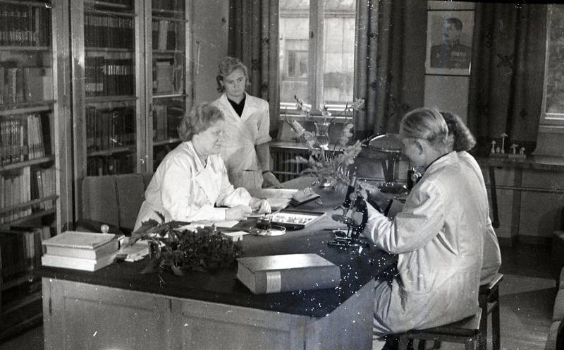 TRÜ farmakognoosiakateedri juhataja (1940-63) prof Alma Tomingas (esiplaanil vasakul) ja TRÜ farmakognoosiakateedri assistent Johannes Tammeorg (esiplaanil paremal) ja kaks naist kirjutuslaua ümber (TRÜ farmaatsiakateeder koos Moskva filmimeestega)