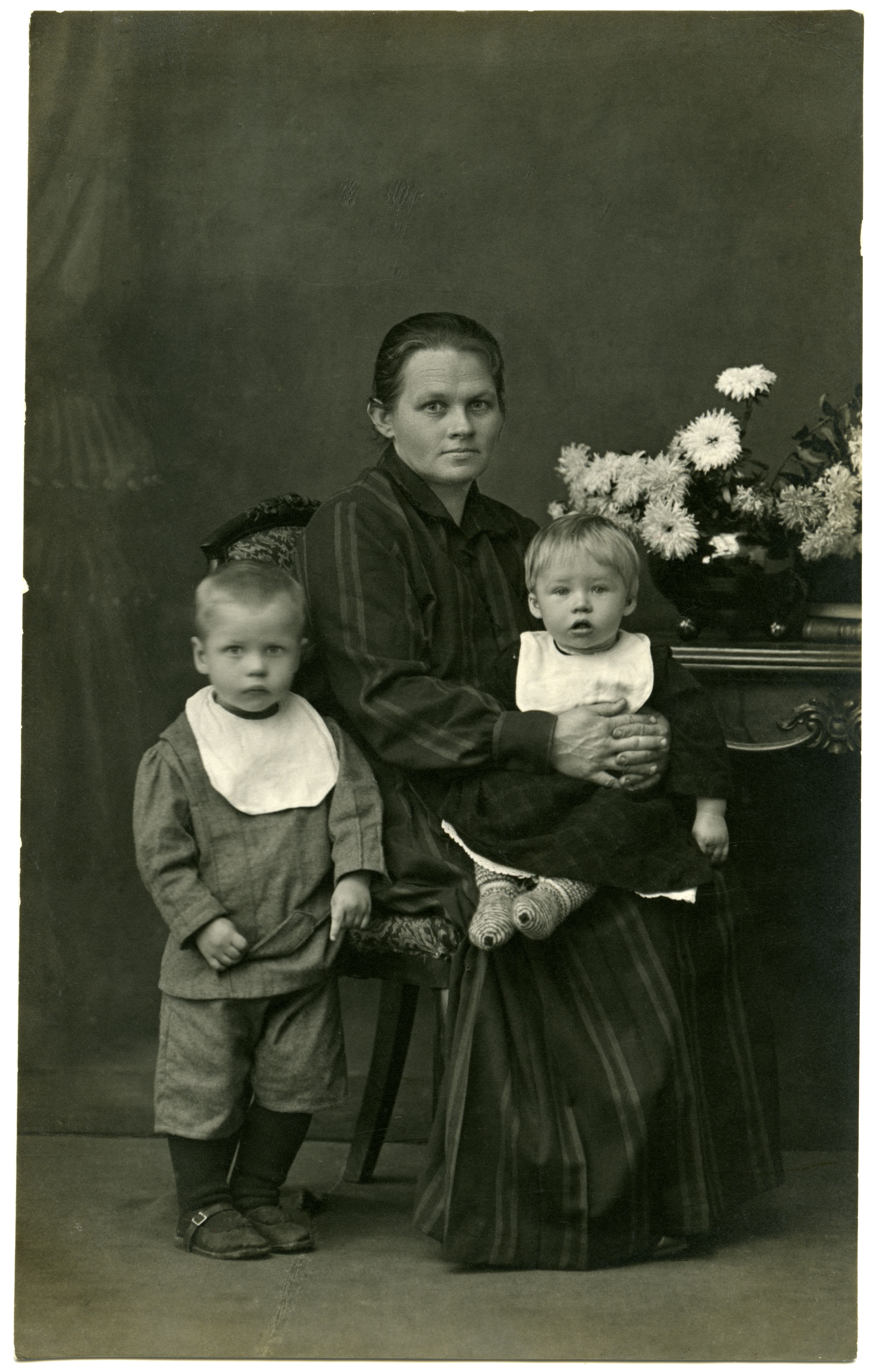 Ema kahe väikelapsega. Emal triibuline kleit, istub väiksem laps süles, poisike seisab tooli juures
