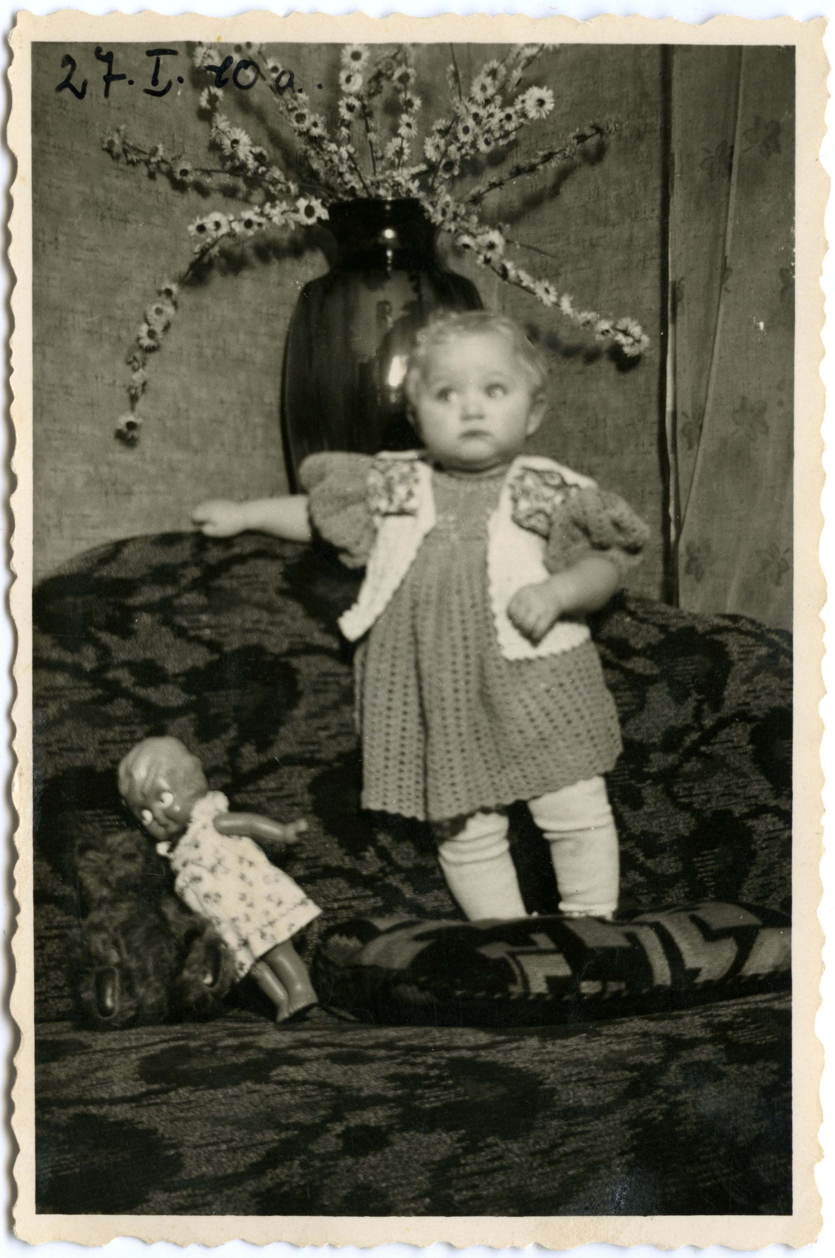 Väikelaps nukuga. Umbes 1 aastane väikelaps kootud kleidis kootud vestiga, seismas diivanil, tema jalgade juures nukk