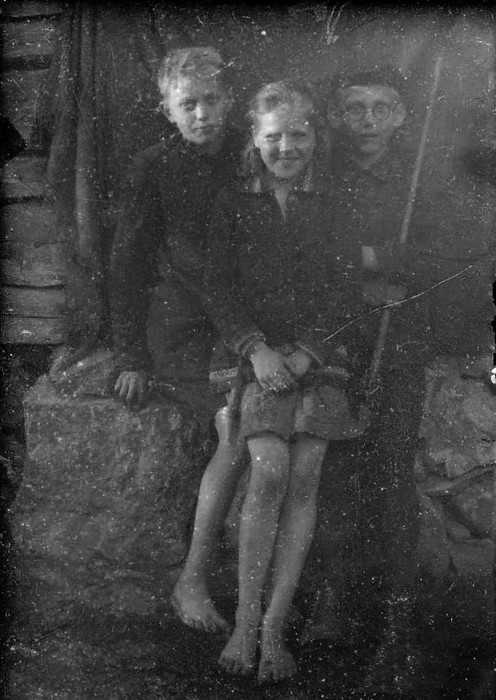 Klaasnegatiiv. Kaks poissi ja tüdruk paljajalu maja vundamendil istumas, seinal riie