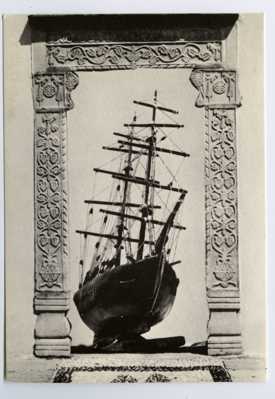 Mykonose Etnograafiamuuseumi postkaart: taimornamentidega ukseraam ja selles 2-mastilise purjelaeva mudel