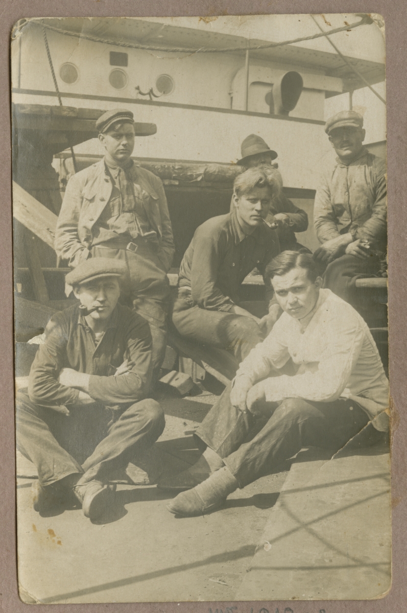 Meremeeste grupifoto: laevatekil istumas kuus tööriides meest, kahel piibud suus