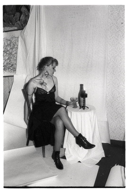 "Vaba teema" -  kehamaalinguga naine joogiklaasi, pudeli ja küünlaga laua ääres toolil