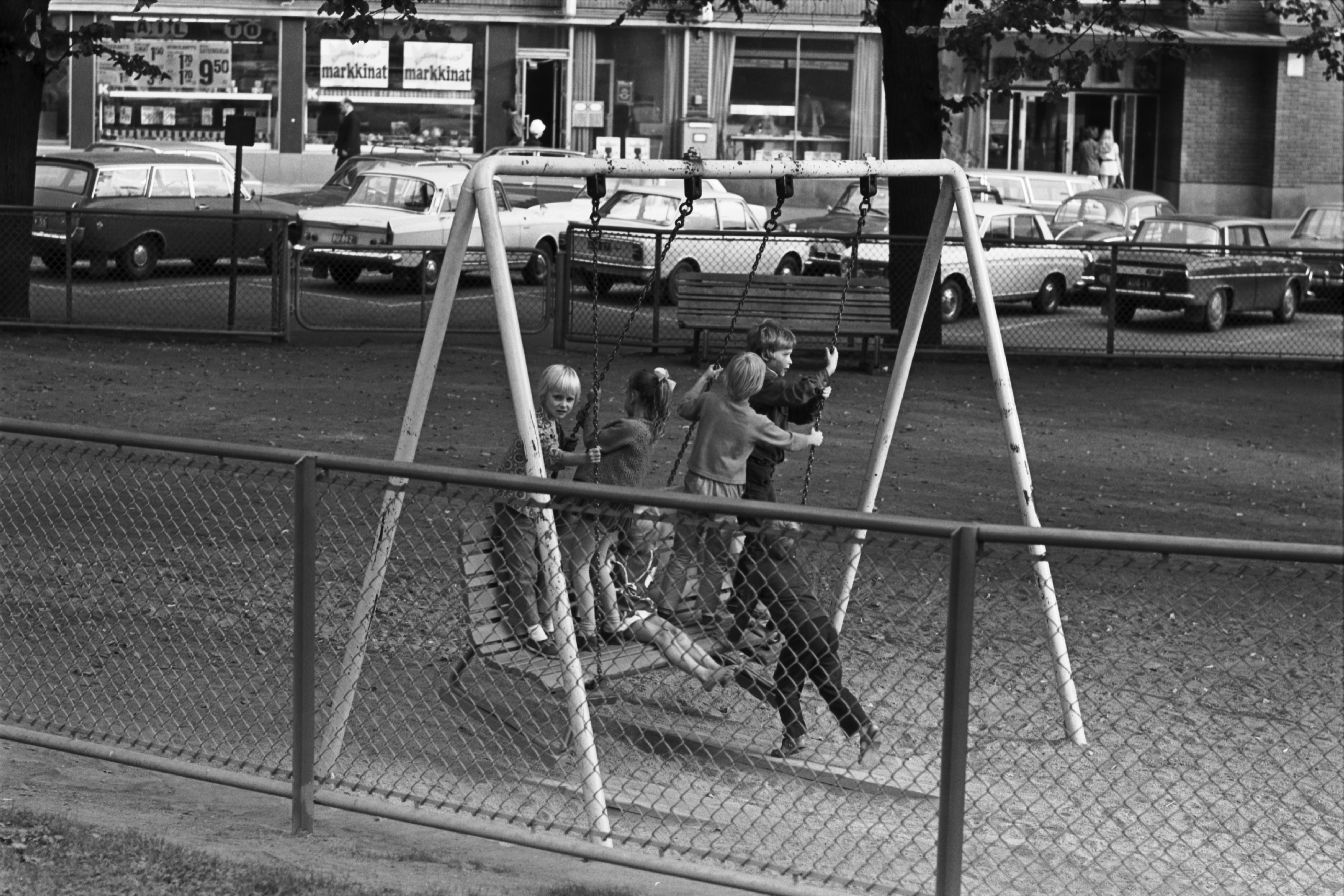 Tove Janssonin puisto. Lapset keinumassa Katajanokan puistossa keinussa, johon on kahden keinulaudan varaan nostettu puiston penkki.