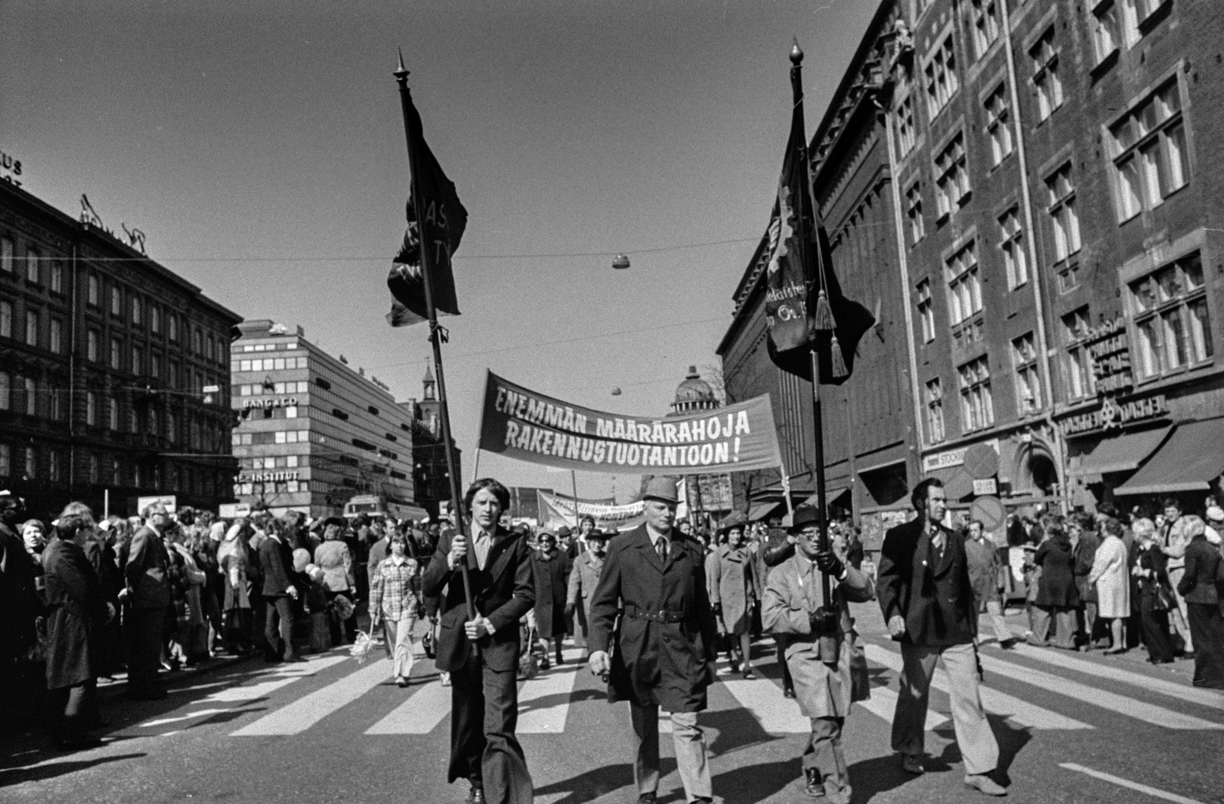 Vappumarssijoita Mannerheimintiellä Stockmannin tavaratalon kohdalla. Marssijat kantavat banderollia, jossa teksti: Enemmän määrärahoja rakennustuotantoon. Kulkueen edessä lipunkantajia.