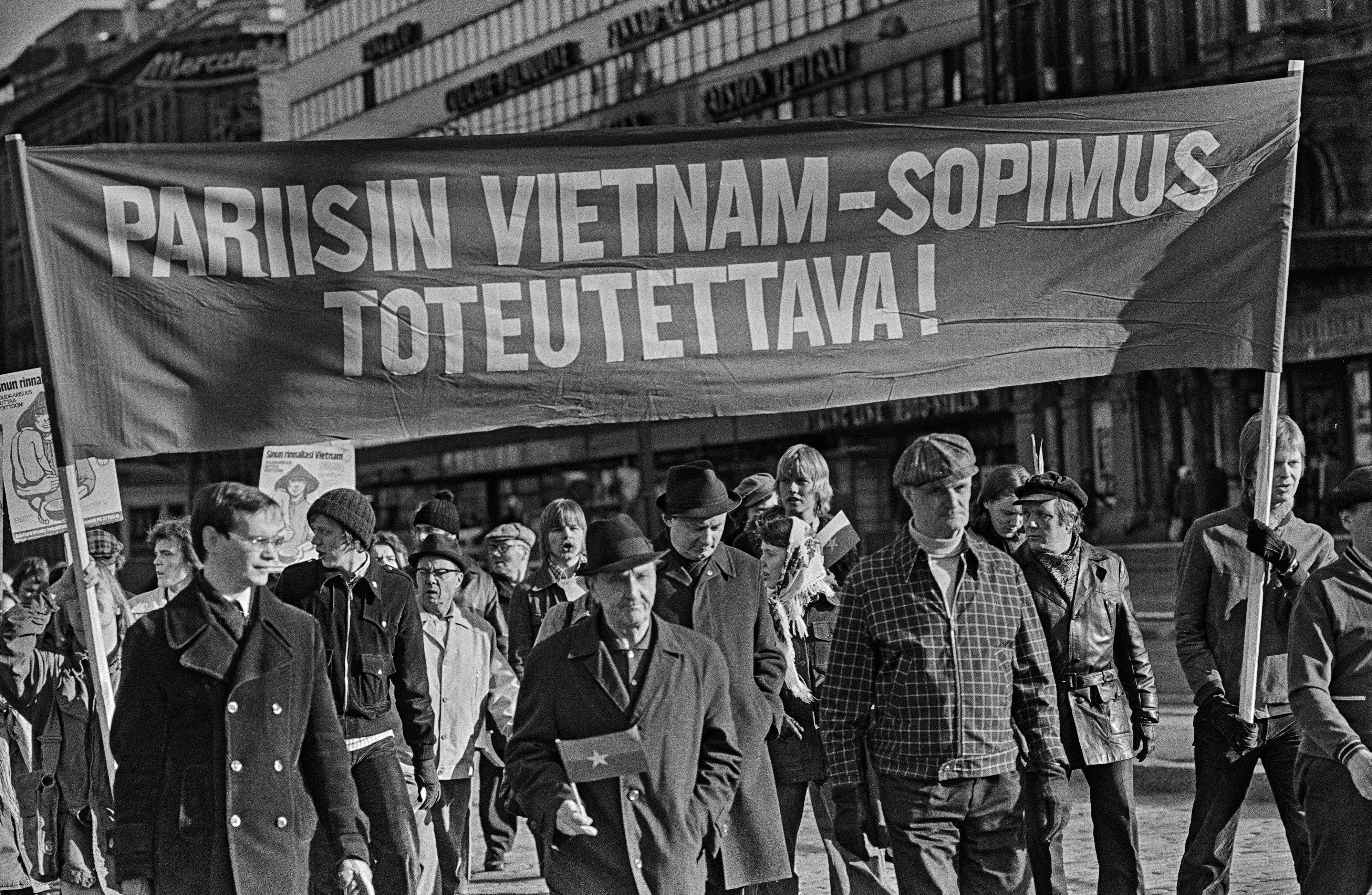 Mielenosoittajia marssimassa ajoradalla Mannerheimintiellä. Mielenosoittajat kantavat banderollia, jossa teksti: Pariisin Vietnam-sopimus toteutettava! Kahdella mielenosoittajalla on kädessään Etelä-Vietnamin kansallisen vapautusrintaman pienoislippu.
