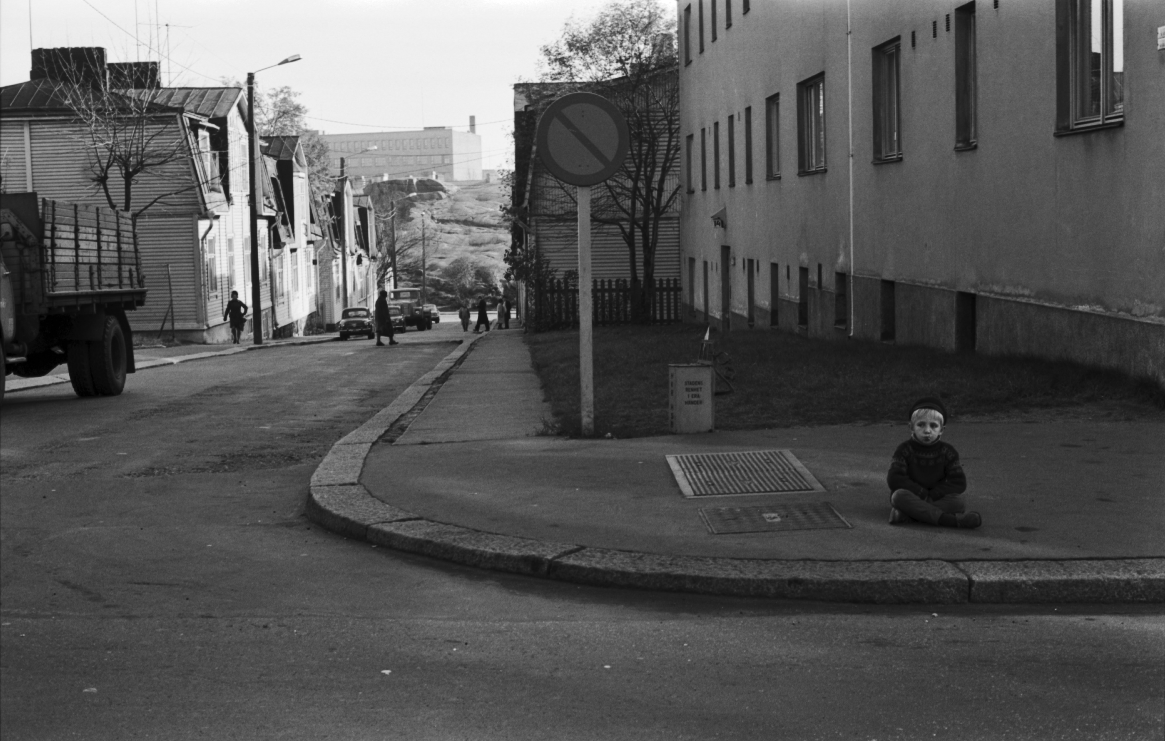 Pieni poika istumassa jalkakäytävällä jalankulkijoiden kulkiessa ohi Sturenkadun ja Virtaintien kulmassa. Näkymä Sturenkadulta Suvannontien suuntaan.