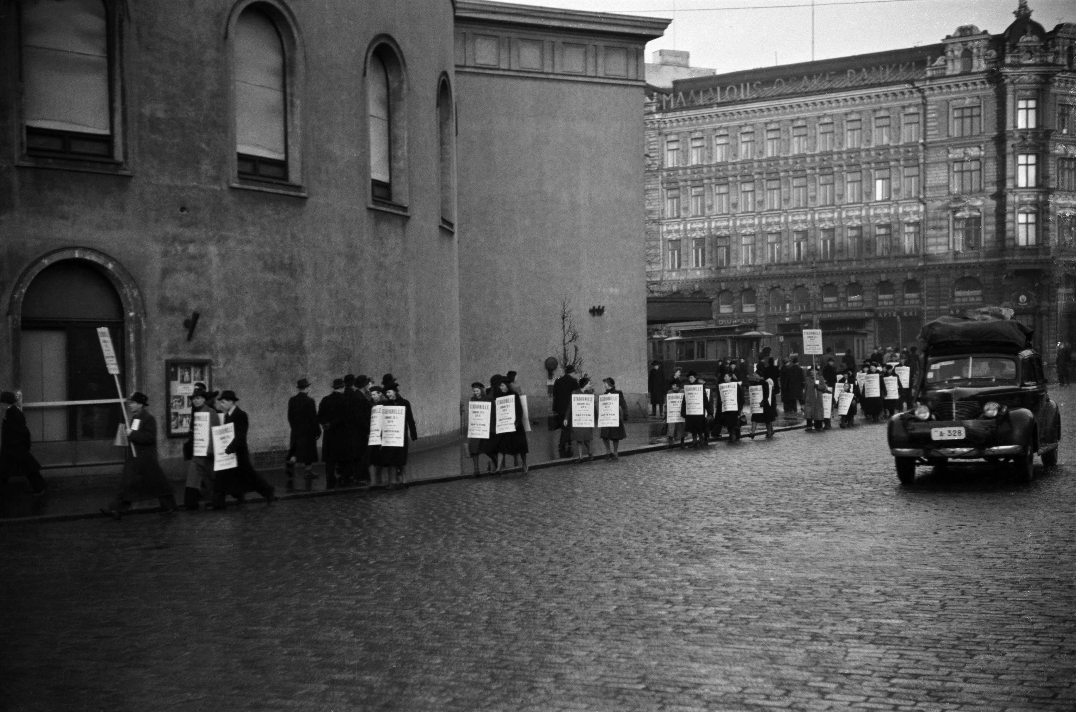 Jehovan todistaja, insinööri Kaarlo Hartevan esitelmää mainostava kulkue Ruotsalaisen teatterin edessä. Taustalla Eteläinen Esplanaadikatu (=Eteläesplanadi) 24. Naisia ja miehiä mainoskylttien kanssa. Oikealla henkilöauto, Chevrolet vm. 1939.