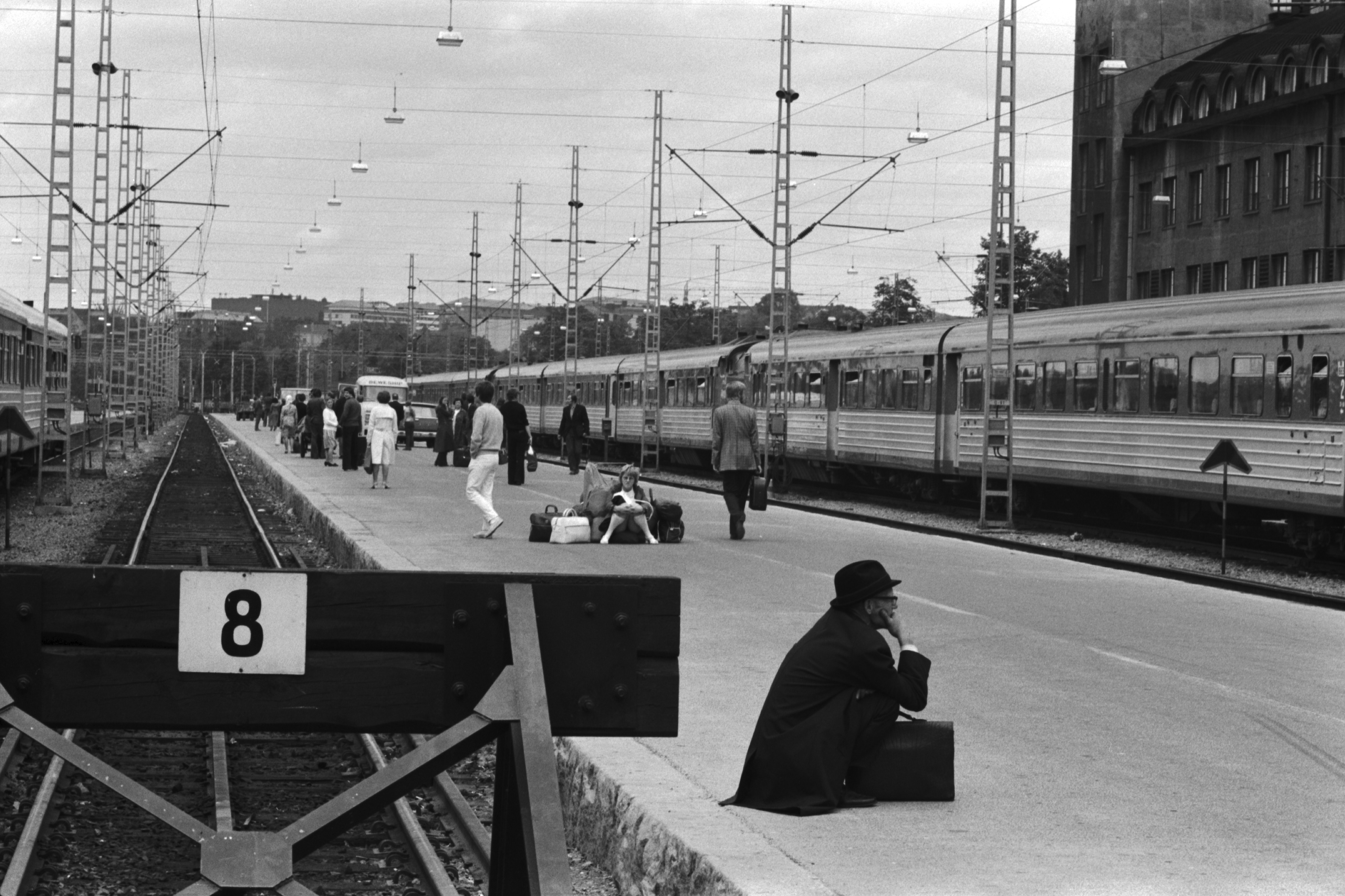 Helsingin rautatieasema. Näkymä raide 8:n päästä pohjoiseen, junien tulosuuntaan. Asemalaiturilla junaa odottavia ihmisiä. Edessä kyykyssä oleva mies salkkuineen, keskellä nuori nainen istumassa suuren matkatavarakasan päällä. Taaempana dieselmoottorijuna.