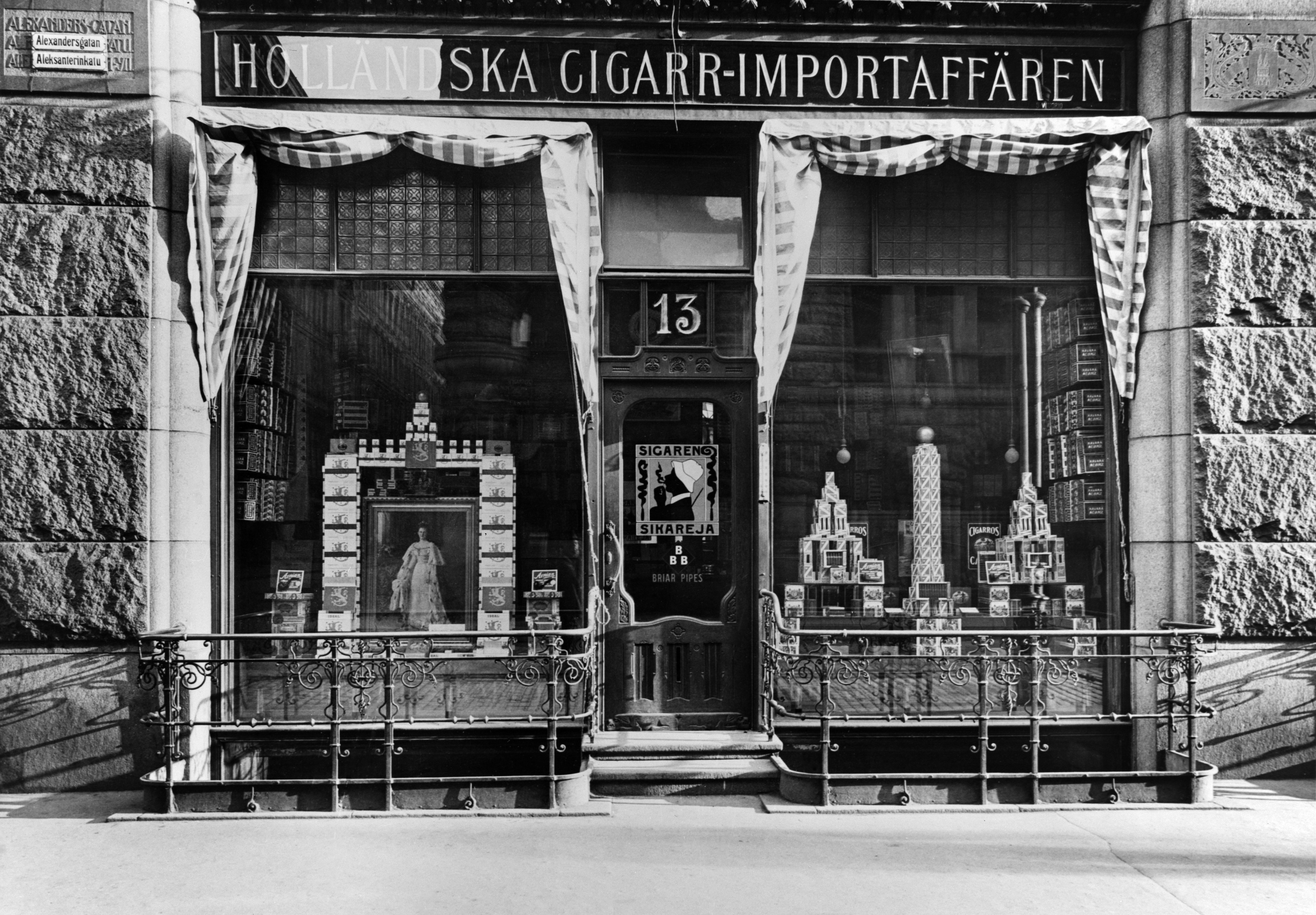 Holländska Cigarrimportaffären Aleksanterinkadulla. Talon seinässä kolmekielinen kadunnimikyltti, jonka päälle on kiinnitetty uusi kaksikielinen kyltti. Holländska Cigarrimportaffärenillä oli v. 1917 kolme liikettä Helsingissä osoitteissa Pohjois-Esplanadi 29, Pohjois-Esplanadi 33 sekä Aleksanterinkatu 13.