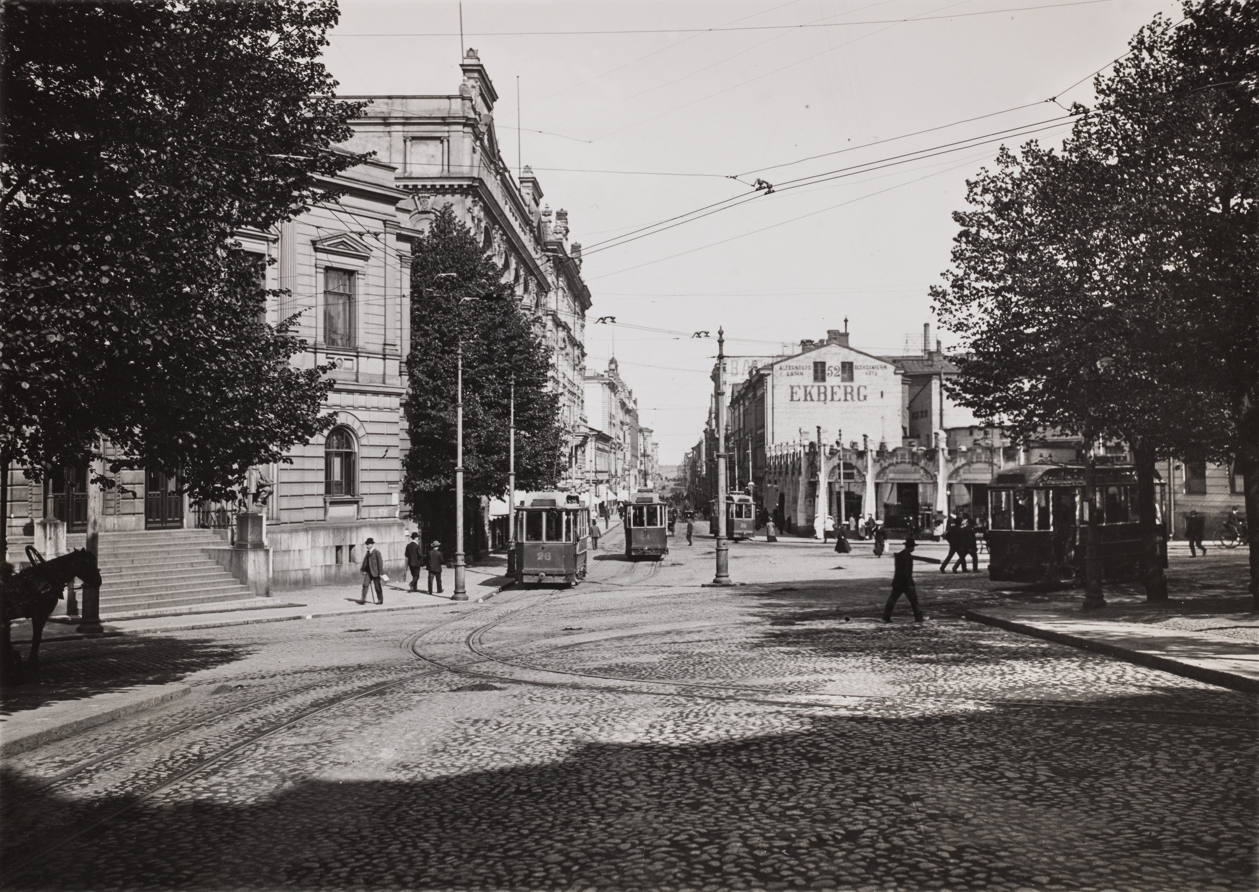 Aleksanterinkadun päätä nykyiseltä Mannerheimintieltä kuvattuna. Vasemmassa reunassa on Vanha Ylioppilastalo. Näkymässä on kaupunkielämää raitiovaunuineen ja hevosajoneuvoineen 1905-vuoden tienoilta. Oikealla olevan kerrostalon seinässä mainostetaan Ekbergin kahvilaa osoitteessa Aleksanterinkatu 52.
