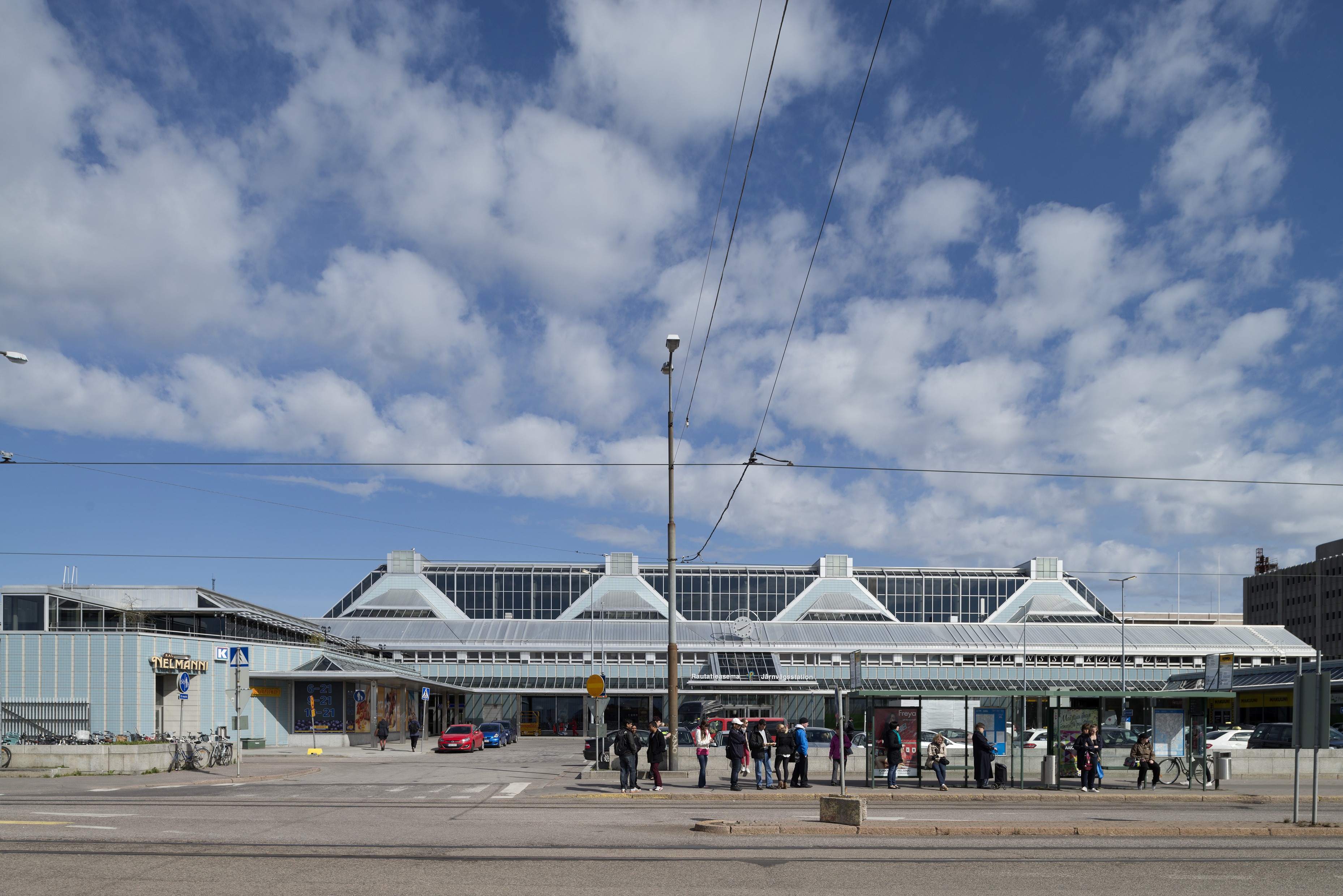 Pasilan rautatieasema Keski-Pasilassa. Arkkitehtitoimisto Jauhiainen - Nuuttila 1991. Teräsrakenteiset laiturikatokset muodostavat ilmavan, läpikuljettavan sillan ratapihan yli. Sininen, nauhamainen muuri osoittaa pääkerroksen tason. Kuvasuunta etelästä Asemapäällikönkadulta, etualalla bussipysäkki ja ihmisiä pysäkillä. Vasemmalla ravintola Nelmanni ja K-market.