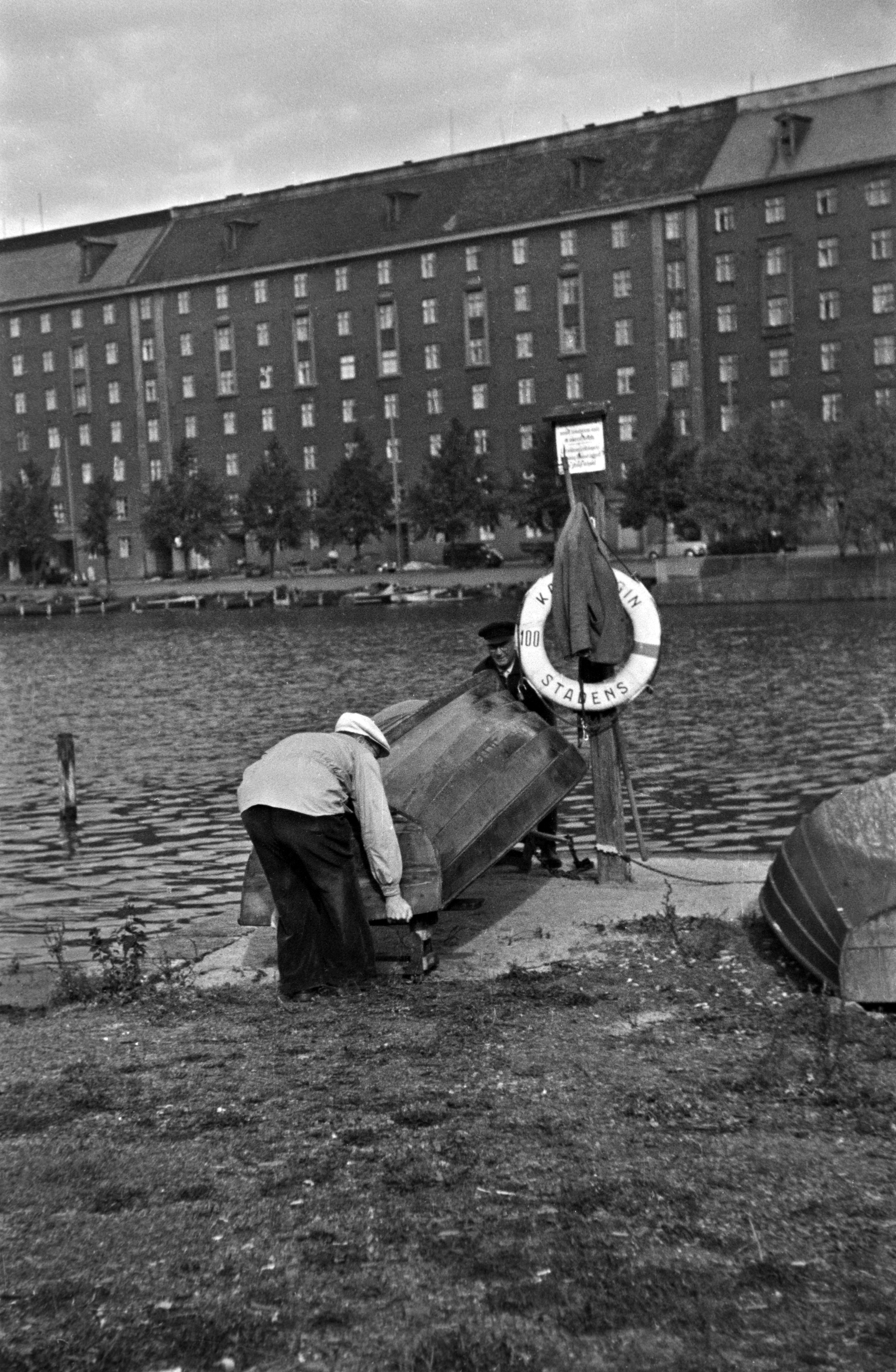 Kaksi miestä nostaa venettä Kaisaniemenlahden rannalla. Taustalla Pitkänsillanranta 5 - 11.