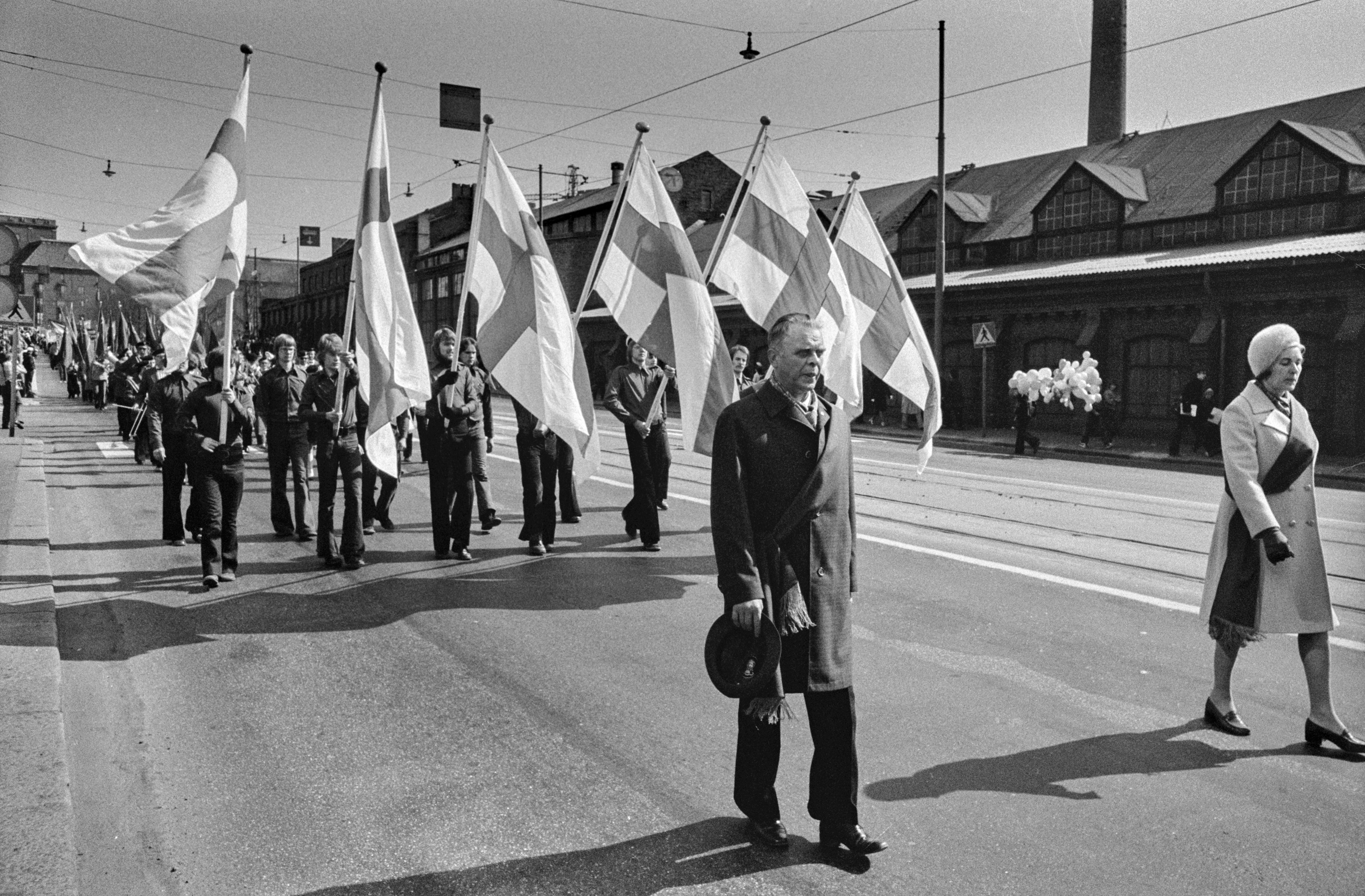 Vappumarssijoita Hämeentiellä. Yhtenäisiin asuihin pukeutuneet nuoret miehet kantavat kulkueen edessä Suomen lippuja.
