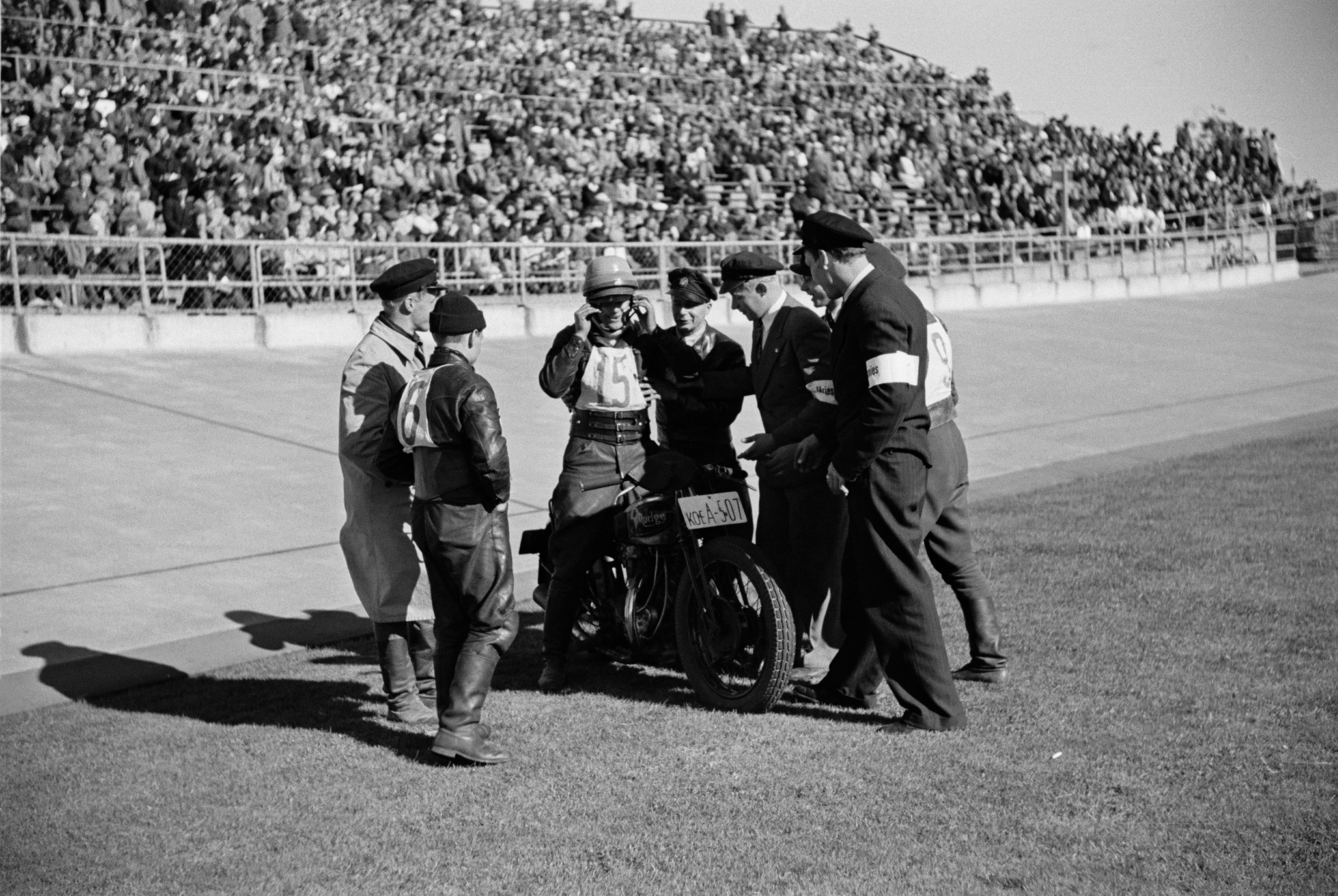 Velodrom, Helsingin pyöräilystadion, moottoripyöräkilpailut. Kilpailija moottoripyörineen radan reunalla, ympärillä toisia kilpailijoita ja järjestysmiehiä.