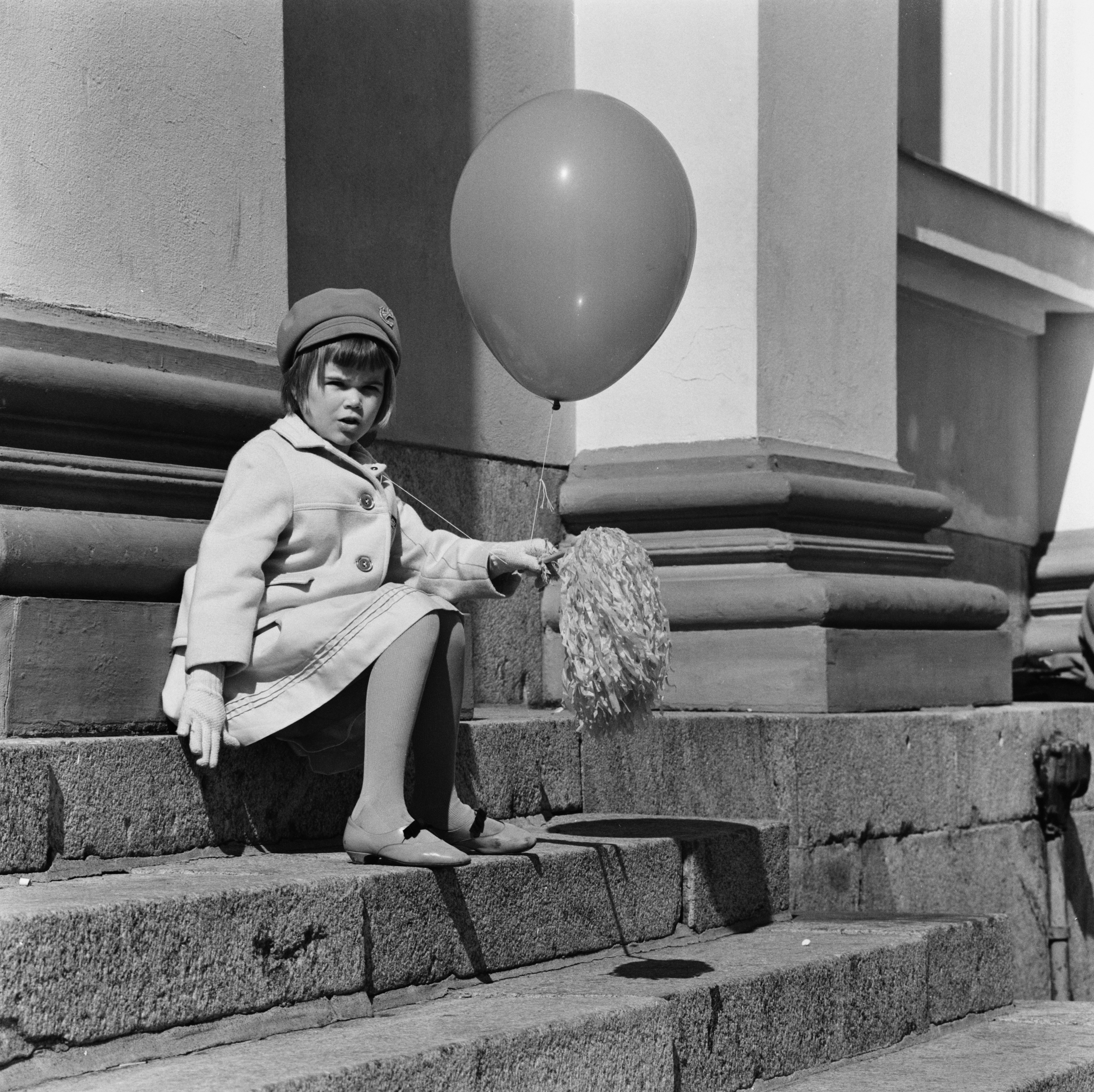 Pikkutyttö istuu vapunpäivänä ilmapallo ja vappuviuhka kädessään Tuomiokirkon portailla..