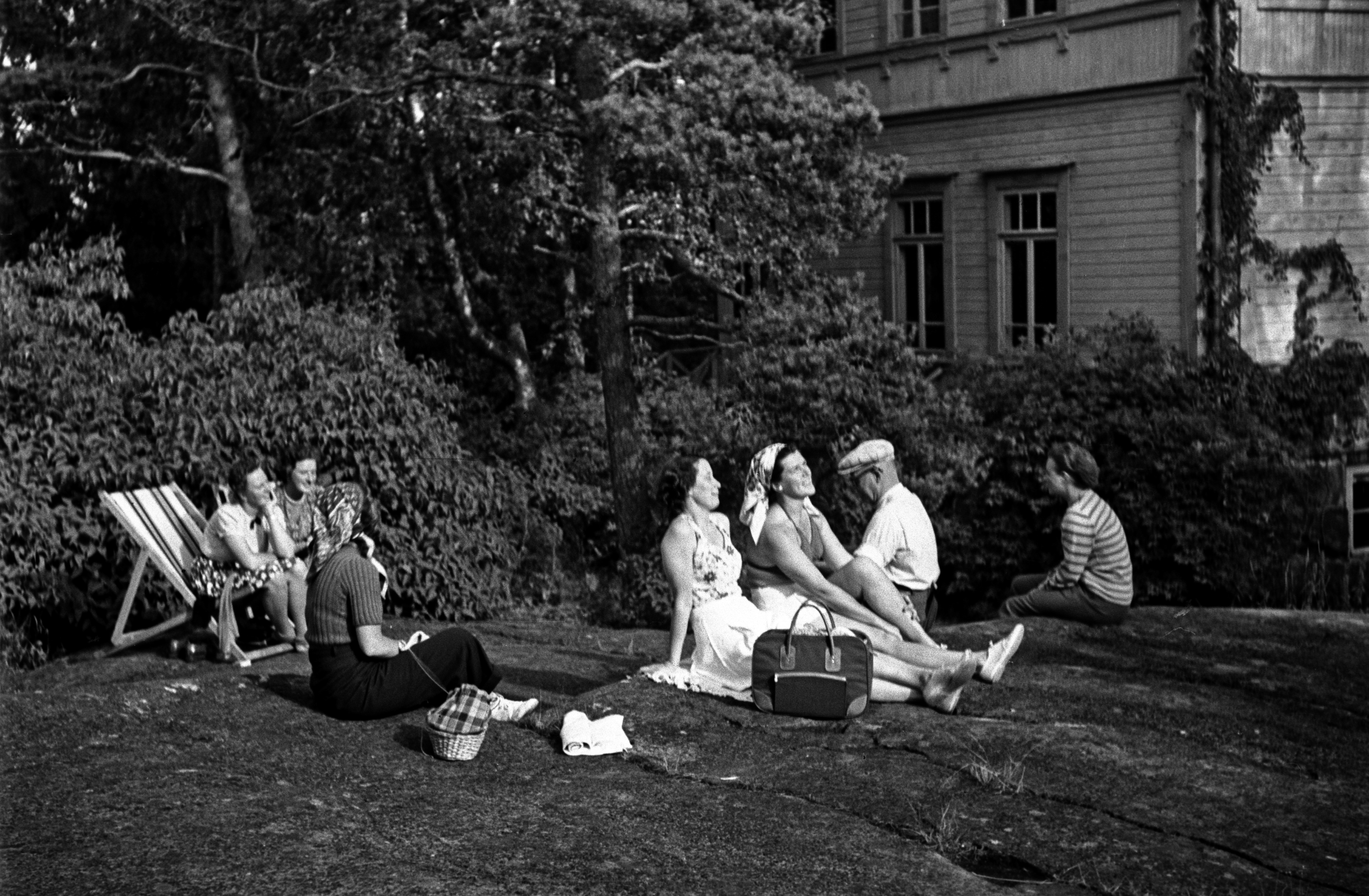 Joukko ihmisiä istumassa ulkona kallioilla ja aurinkotuoleissa Pihlajasaaressa. Oikealla näkyy nk. "Elannon huvila", Wästanhäll.
