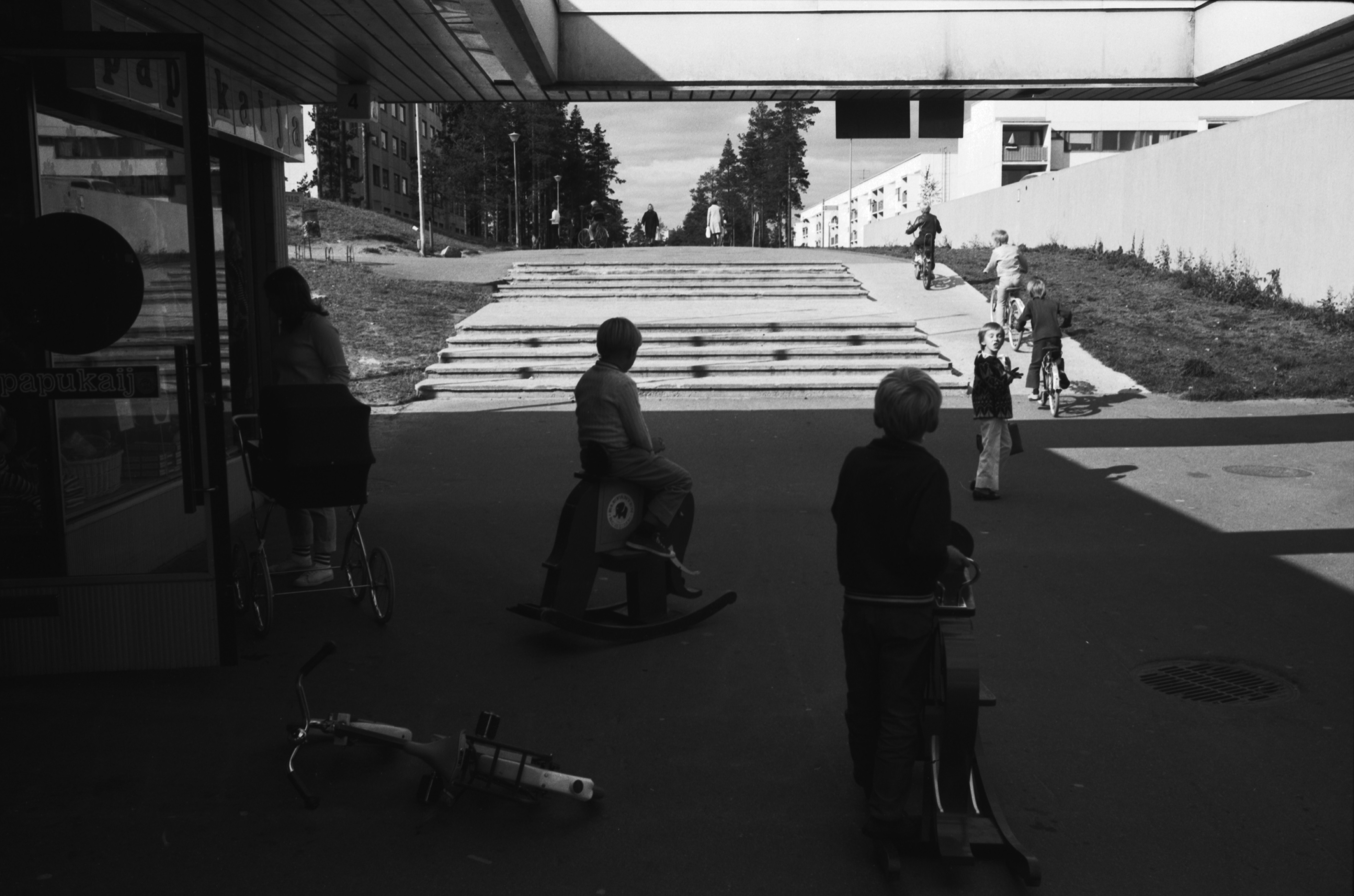 Isännänpolku. Kontulan ostoskeskus. Kontulan ostoskeskukseen johtavat portaat Isännänpolun päässä. Etualalla lapsia polkupyörineen sekä kaksi lasta keinuelefanttien selässä lastenvaateliike Papukaijan edessä.